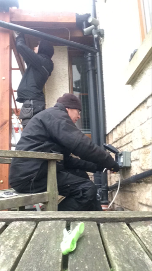 One man in the background standing working with some wiring above his head. One man in the foreground wiring an outside socket on a house wall